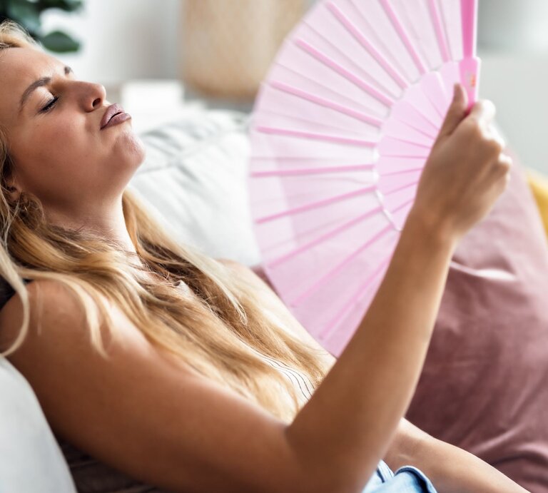 Junge blonde Frau mit müdem Gesichtsausdruck fächelt sich auf dem Sofa mit rosa Fächer Luft zu, sichtbar erschöpft von Hitze. | © Adobe Stock / nenetus