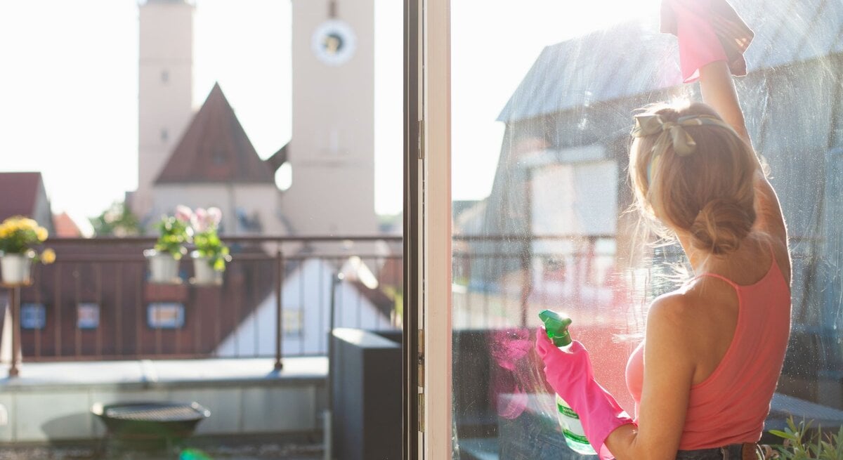  Eine Frau mit aufgesteckten Haaren und rosafarbenem Oberteil steht auf einem Balkon und putzt das Fensterglas mit einem Sprühreiniger und einem Tuch. Im Hintergrund sieht man verschwommen einen Kirchturm und ein weiteres Gebäude unter einem blauen Himmel. Sie trägt rosa Gummihandschuhe und ist im Profil sichtbar, während sie sich auf die Reinigungsarbeit konzentriert.