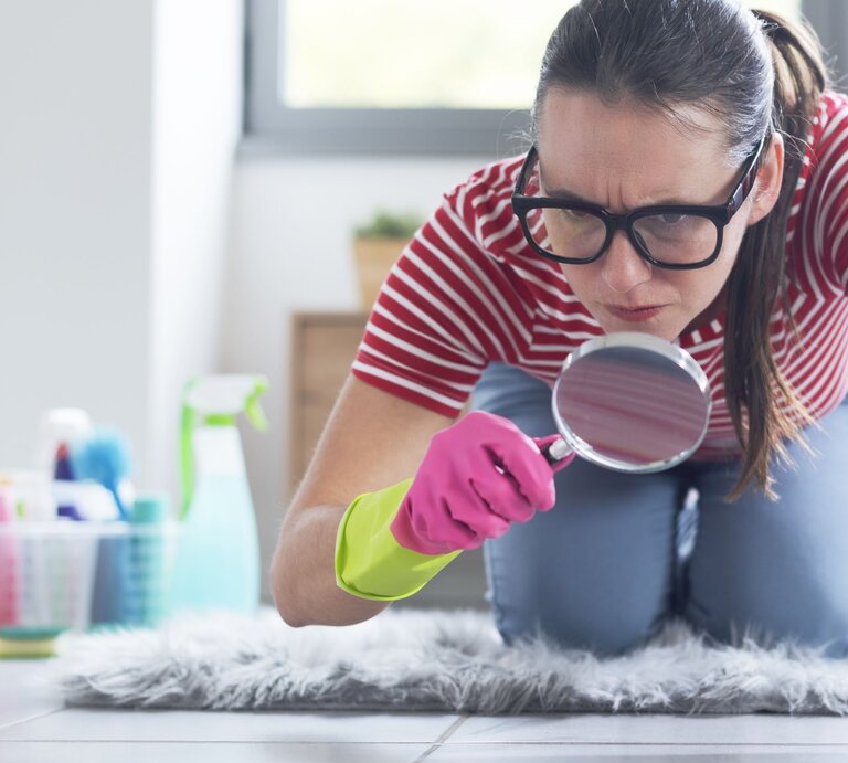Junge Frau mit Brille und Putzhandschuhen kniet ernsthaft auf dem Boden und sucht mit Lupe nach Schmutz im Badezimmer. | © Adobe Stock / stokkete