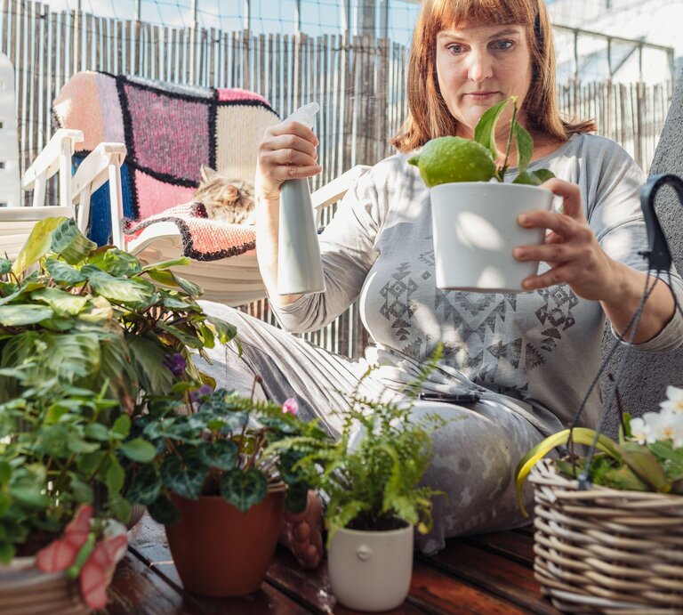 Eine rothaarige Frau in grauem Pyjama pflegt Pflanzen auf einem Balkon, im Hintergrund liegt eine Katze auf einem Stuhl. | © Adobe Stock / larisikstefania