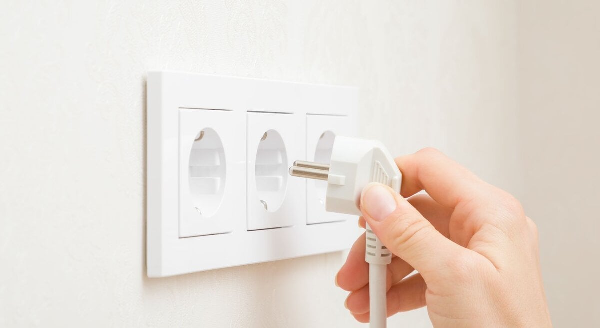 Eine Hand steckt einen weißen Stecker in eine von drei weißen Steckdosen an einer hellen Wand.