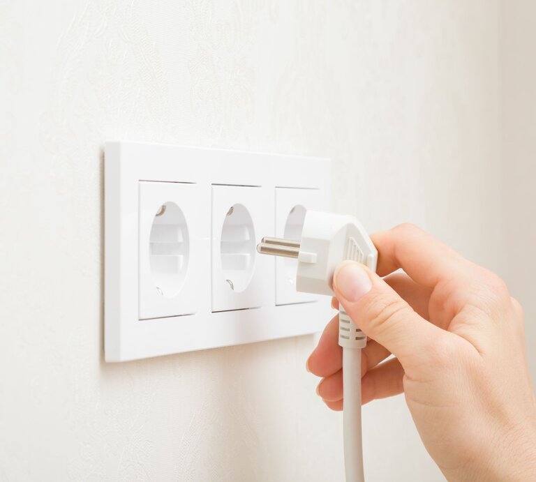 Eine Hand steckt einen weißen Stecker in eine von drei weißen Steckdosen an einer hellen Wand. | © Adobe Stock/fotoduets