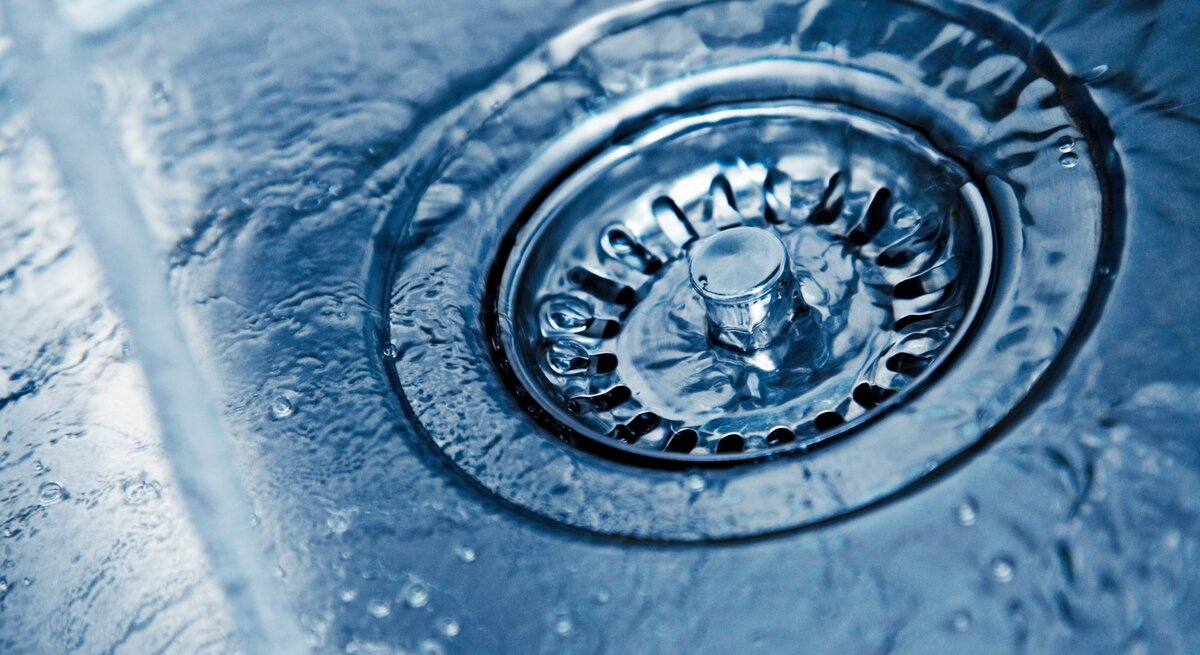 Nahaufnahme eines glänzenden Abflusses in einem Edelstahlspülbecken mit fließendem Wasser und spiegelnden Wassertropfen.