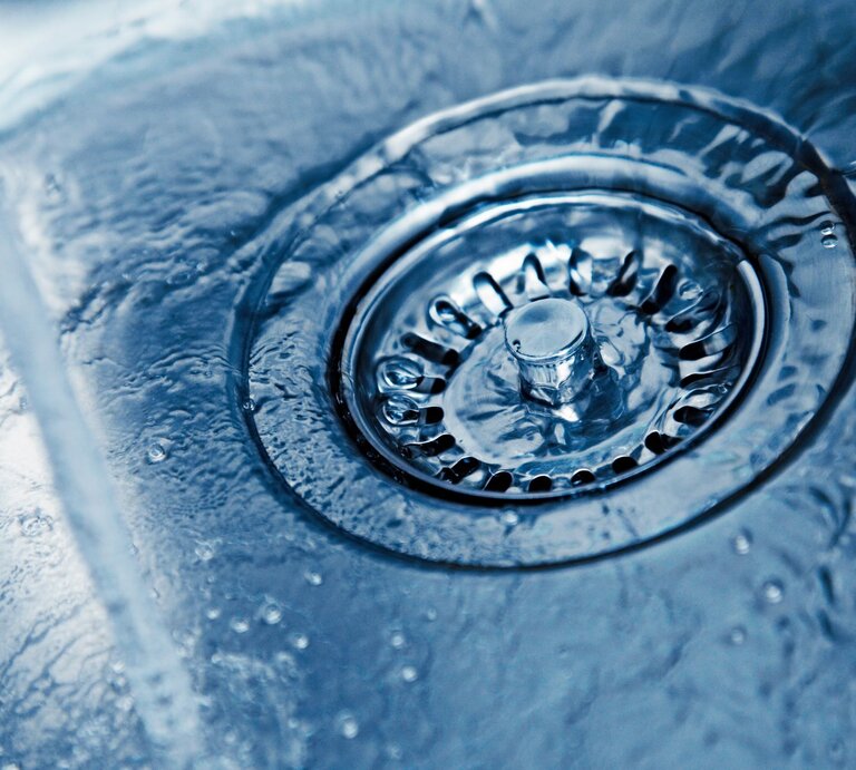 Nahaufnahme eines glänzenden Abflusses in einem Edelstahlspülbecken mit fließendem Wasser und spiegelnden Wassertropfen. | © Adobe Stock/Friedberg