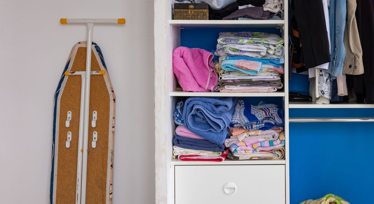 Ein geöffnetes Bügelbrett, das neben einem weißen Kleiderschrank mit einer blauen Tür steht. Im Schrank sind ordentlich gefaltete Handtücher und Bettwäsche auf Regalen sowie hängende Kleidungsstücke wie Jacken und Hosen zu sehen. Unter den Regalen befinden sich Schubladen, von denen zwei geschlossen sind und eine offen mit mehr Wäsche darin."