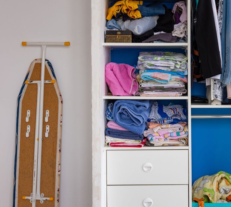 Ein geöffnetes Bügelbrett, das neben einem weißen Kleiderschrank mit einer blauen Tür steht. Im Schrank sind ordentlich gefaltete Handtücher und Bettwäsche auf Regalen sowie hängende Kleidungsstücke wie Jacken und Hosen zu sehen. Unter den Regalen befinden sich Schubladen, von denen zwei geschlossen sind und eine offen mit mehr Wäsche darin." | © Adobe Stock/ madhourse