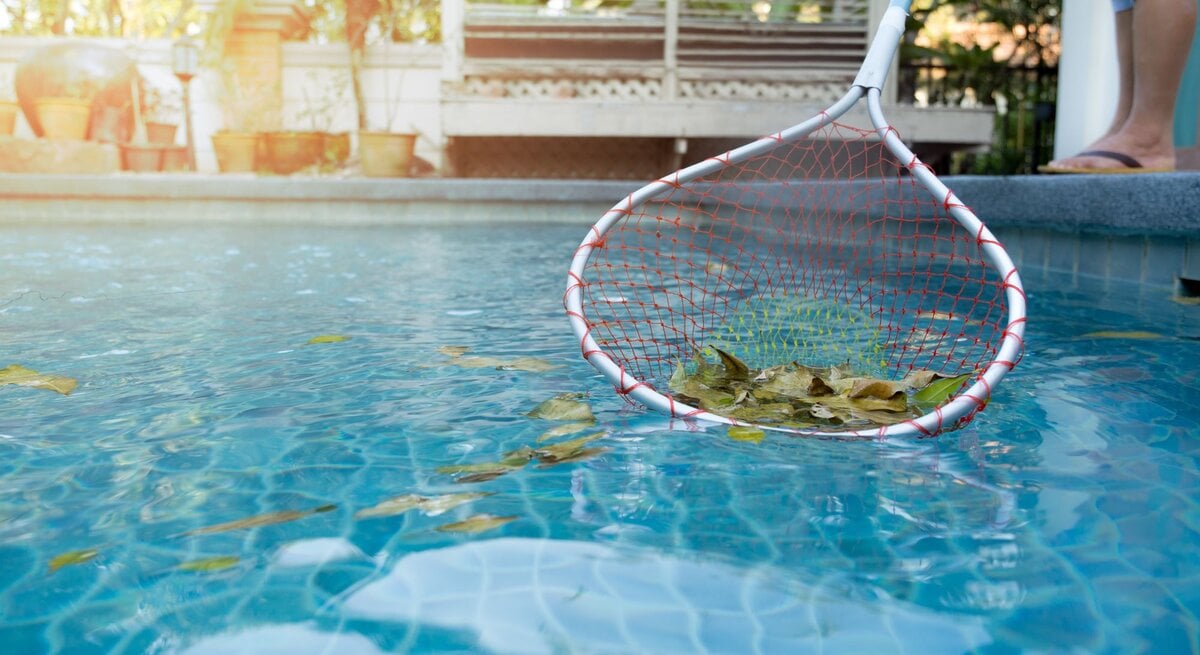 Ein Mensch in Flipflops fischt mit einem Kescher herabgefallene Blätter aus einem blauen Pool bei sanftem Sonnenlicht.