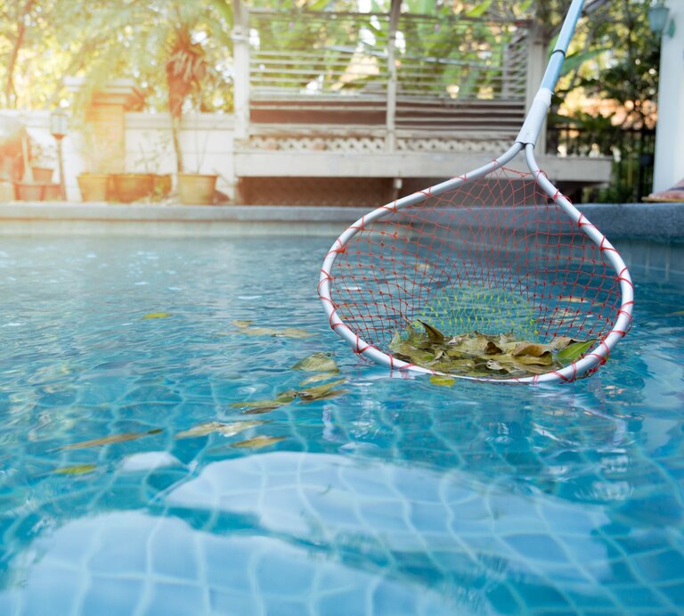 Ein Mensch in Flipflops fischt mit einem Kescher herabgefallene Blätter aus einem blauen Pool bei sanftem Sonnenlicht. | © AdobeStock/bignai