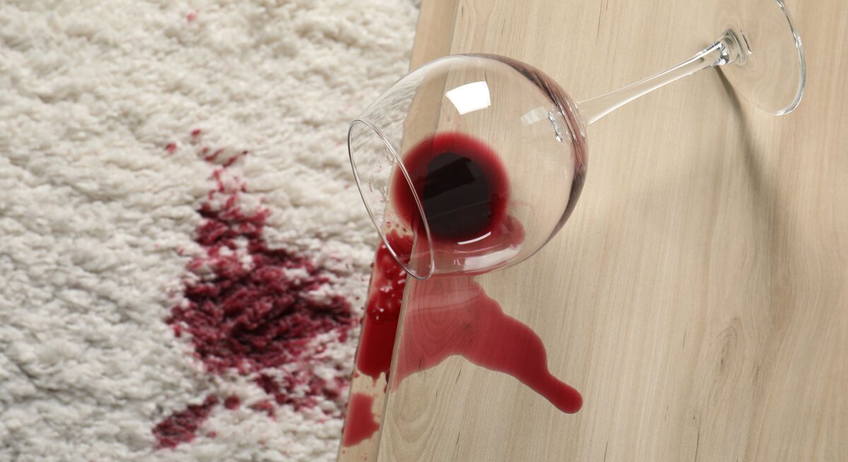 Ein umgekipptes Weinglas auf einer Holzoberfläche, aus dem roter Wein auf einen weißen Teppich mit einem großen Fleck ausgelaufen ist.