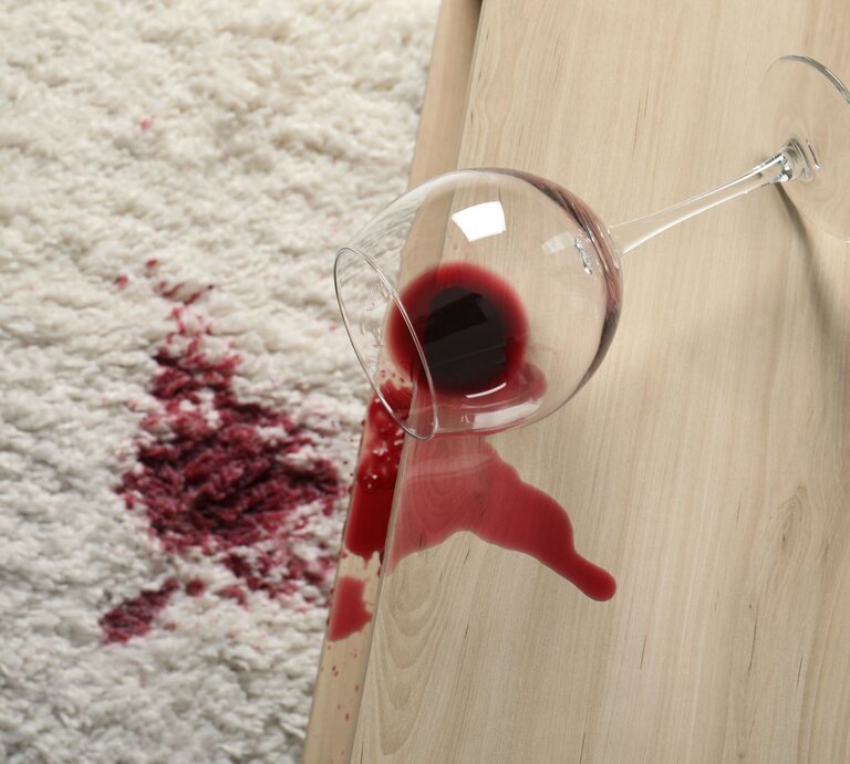 Ein umgekipptes Weinglas auf einer Holzoberfläche, aus dem roter Wein auf einen weißen Teppich mit einem großen Fleck ausgelaufen ist. | © Shutterstock/New Africa