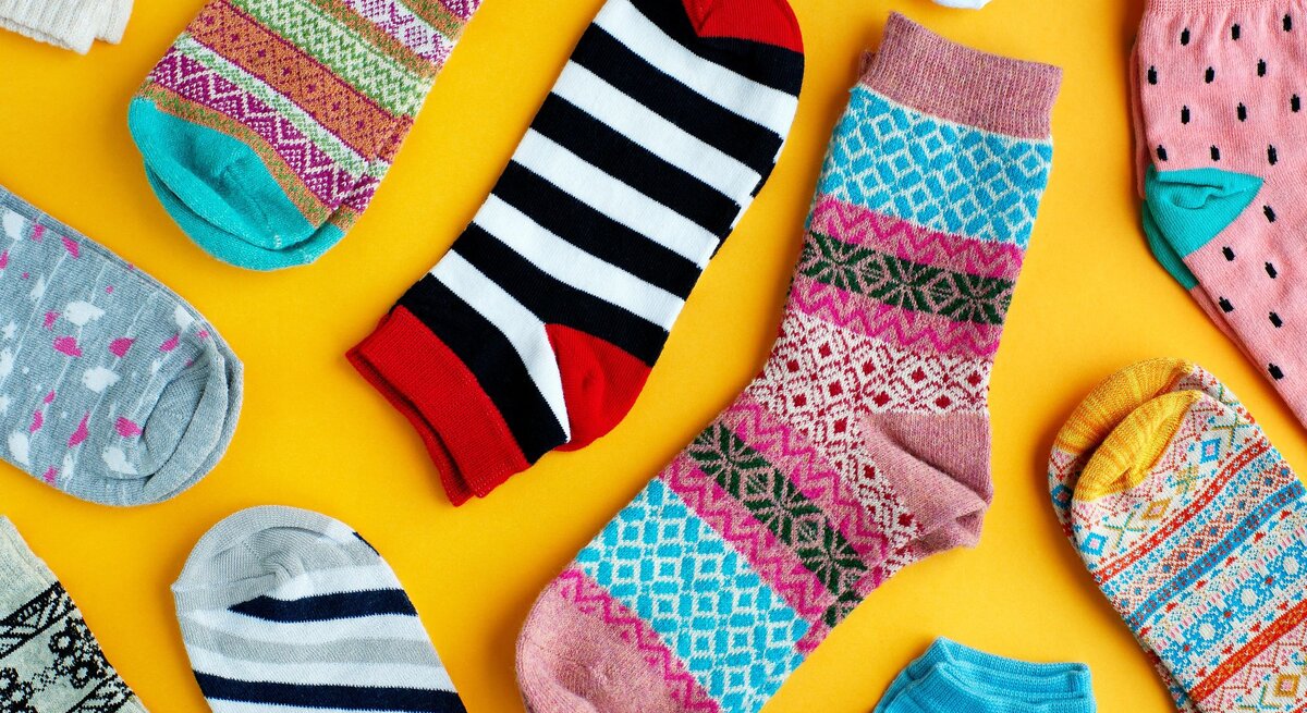 Bunte Auswahl verschiedener Socken auf einem leuchtend gelben Hintergrund. Die Socken weisen vielfältige Muster und Farben auf, darunter Streifen, Punkte, und geometrische Designs in Farben von Rot und Schwarz bis hin zu Pastelltönen.