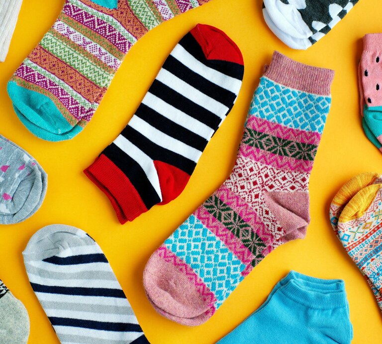 Bunte Auswahl verschiedener Socken auf einem leuchtend gelben Hintergrund. Die Socken weisen vielfältige Muster und Farben auf, darunter Streifen, Punkte, und geometrische Designs in Farben von Rot und Schwarz bis hin zu Pastelltönen. | © Shutterstock/ Evgeniya369
