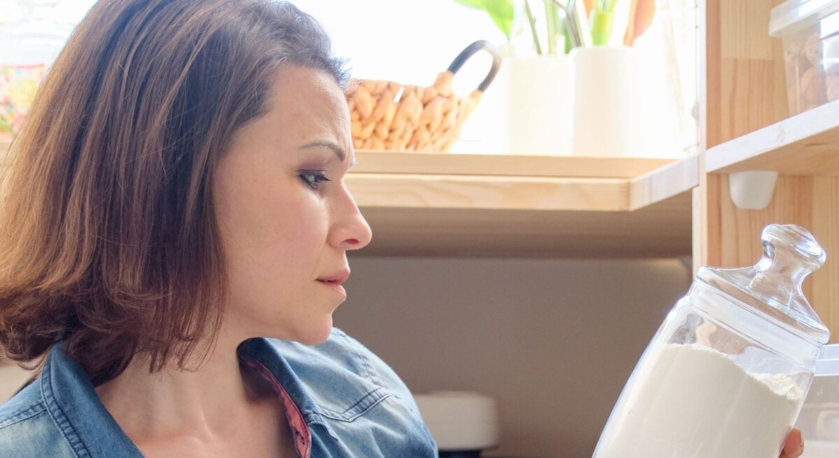 Junge Frau mit kinnlangem braunem Haar schaut nachdenklich auf ein großes Glas mit weißem Mehl in heller Küchenvorratskammer.