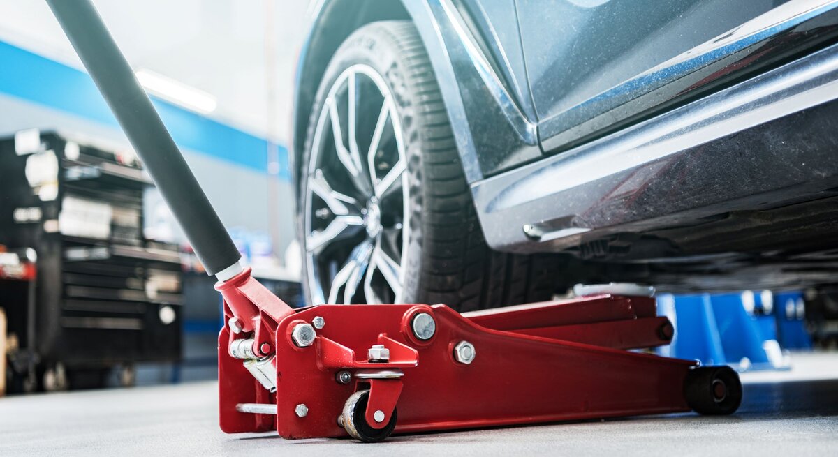 Ein roter Wagenheber hebt ein dunkles Auto in einer hellen Werkstatt an, das Vorderrad mit silberner Felge schwebt über dem Boden.