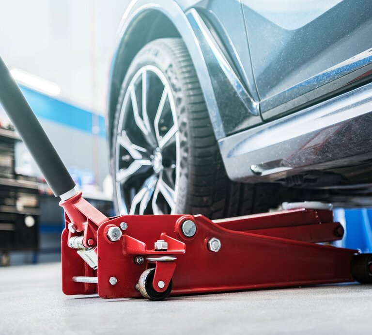 Ein roter Wagenheber hebt ein dunkles Auto in einer hellen Werkstatt an, das Vorderrad mit silberner Felge schwebt über dem Boden. | © Shutterstock/Virrage Images