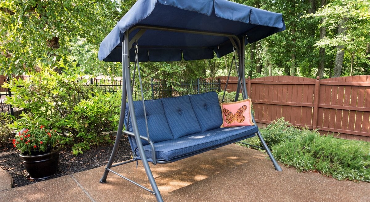  Dunkelblaue Gartenschaukel mit Sonnendach und Polstern steht auf Terrasse, umgeben von grünen Sträuchern und Holzzaun.