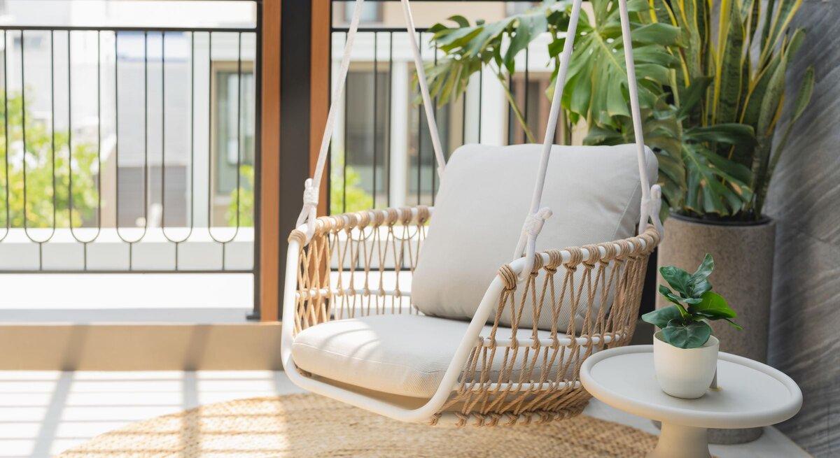   Heller Hängesessel mit beigem Kissen aus Seilgeflecht steht auf sonnigem Balkon neben Pflanzen und kleinem Beistelltisch.