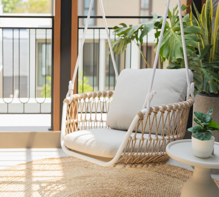   Heller Hängesessel mit beigem Kissen aus Seilgeflecht steht auf sonnigem Balkon neben Pflanzen und kleinem Beistelltisch. | © GettyImages/whyframestudio
