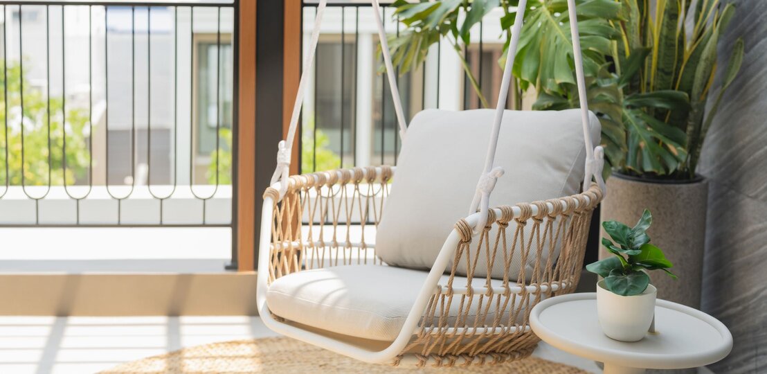   Heller Hängesessel mit beigem Kissen aus Seilgeflecht steht auf sonnigem Balkon neben Pflanzen und kleinem Beistelltisch. | © GettyImages/whyframestudio