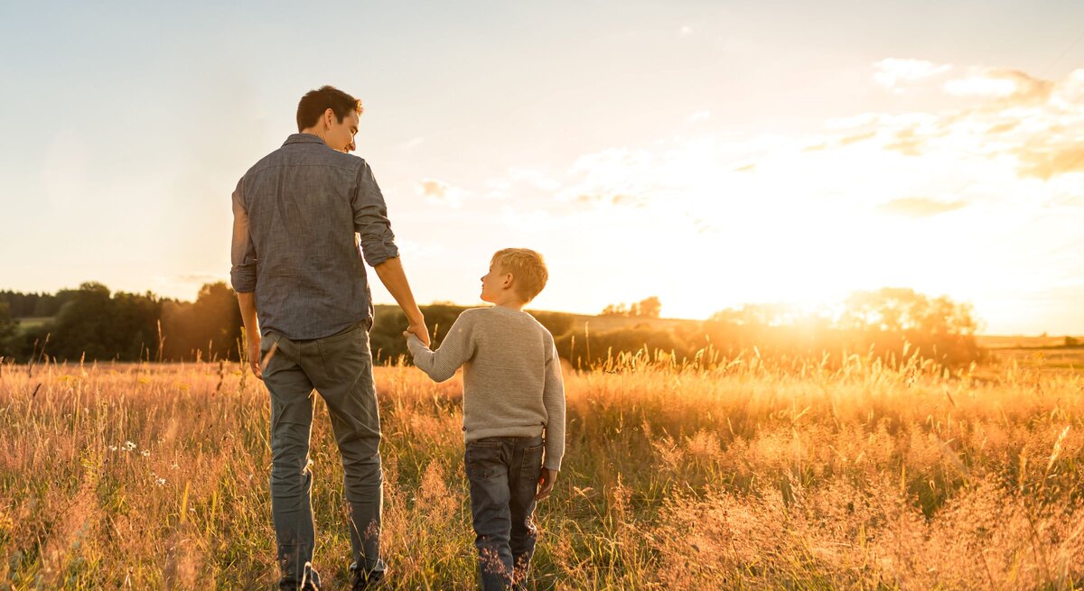 Junger Mann im blauen Hemd geht bei Sonnenuntergang durch hohes Gras und hält die Hand eines blonden Jungen, beide lächeln.