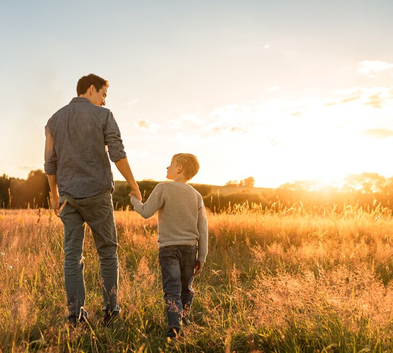 Junger Mann im blauen Hemd geht bei Sonnenuntergang durch hohes Gras und hält die Hand eines blonden Jungen, beide lächeln. | © gettyImages/	kieferpix