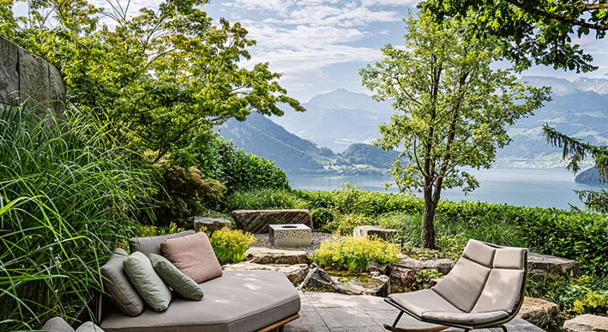 Moderne Terrasse mit Lounge und Schaukelstuhl zwischen viel Grün, Blick auf einen See und umliegende Berglandschaft unter locker bewölktem Himmel.