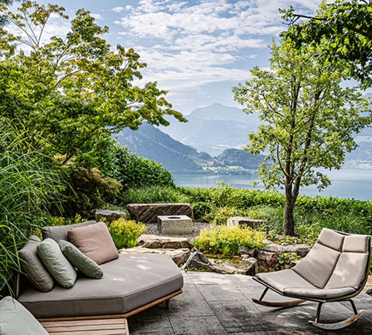 Moderne Terrasse mit Lounge und Schaukelstuhl zwischen viel Grün, Blick auf einen See und umliegende Berglandschaft unter locker bewölktem Himmel. | © Callwey Verlag/ Fer­di­nand Graf Luck­ner
