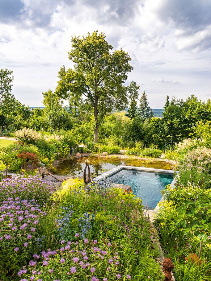 ppiger Garten mit blühenden Stauden im Vordergrund, daneben ein rechteckiger Naturpool mit Holzsteg und Skulptur; dahinter dichter Grünbewuchs und ein großer Baum unter wolkigem Himmel. | © Callwey Verlag/ Syl­via Knit­tel