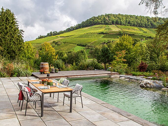 Steinterrasse mit gedecktem Esstisch am Naturpool, dahinter Weinberge und sanfte Hügel unter wechselhaftem Himmel. | © Callwey Verlag/ Apostolos Simeonakis