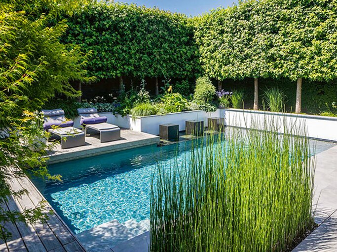 Moderner Gartenpool mit klarem Wasser, gesäumt von Holz- und Steinterrassen, Lounge-Möbeln und Hochbeeten; dichte grüne Hecken und geschnittene Bäume bieten Sichtschutz. | © Callwey Verlag/ Tom Bendix