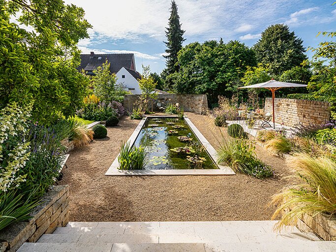 Formaler Innenhofgarten mit langem rechteckigen Wasserbecken, Kiesflächen, Natursteinmauern und Sitzplatz unter Sonnenschirm. | © Callwey Verlag/ Thorsten Scherz