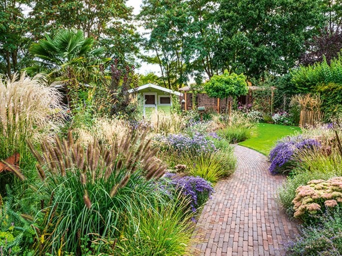 Geschwungener Klinkerweg führt durch hohe Ziergräser und spätblühende Stauden zu einem kleinen Gartenhaus. | © Callwey Verlag/ Ulrike Romeis