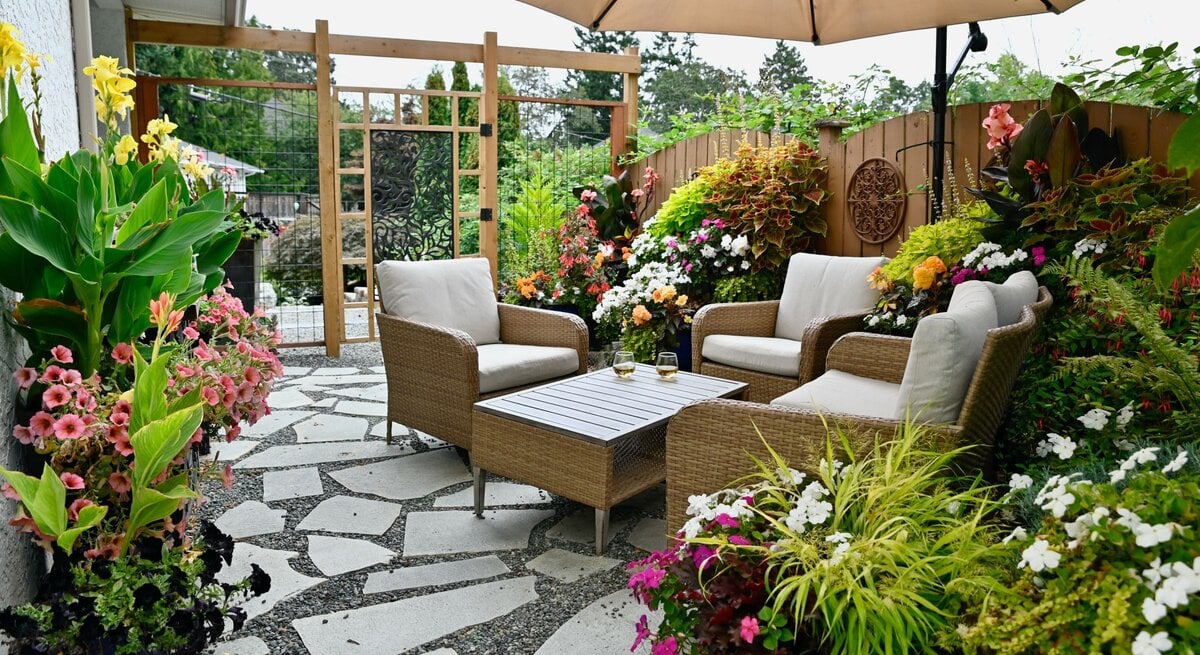 Gemütliche Gartenterrasse mit Korbsesseln unter Sonnenschirm, umgeben von üppigen Blumen und hellem Steinboden. | © Shutterstock/ Klem Mitch