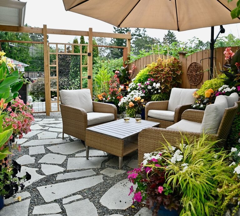 Gemütliche Gartenterrasse mit Korbsesseln unter Sonnenschirm, umgeben von üppigen Blumen und hellem Steinboden. | © Shutterstock/ Klem Mitch