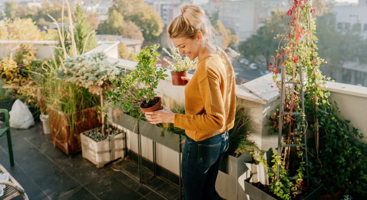 Eine Frau beim Urban Gardening auf ihrem Balkon mit Blumentöpfen und Gemüsepflanzen am Geländer und am Boden.