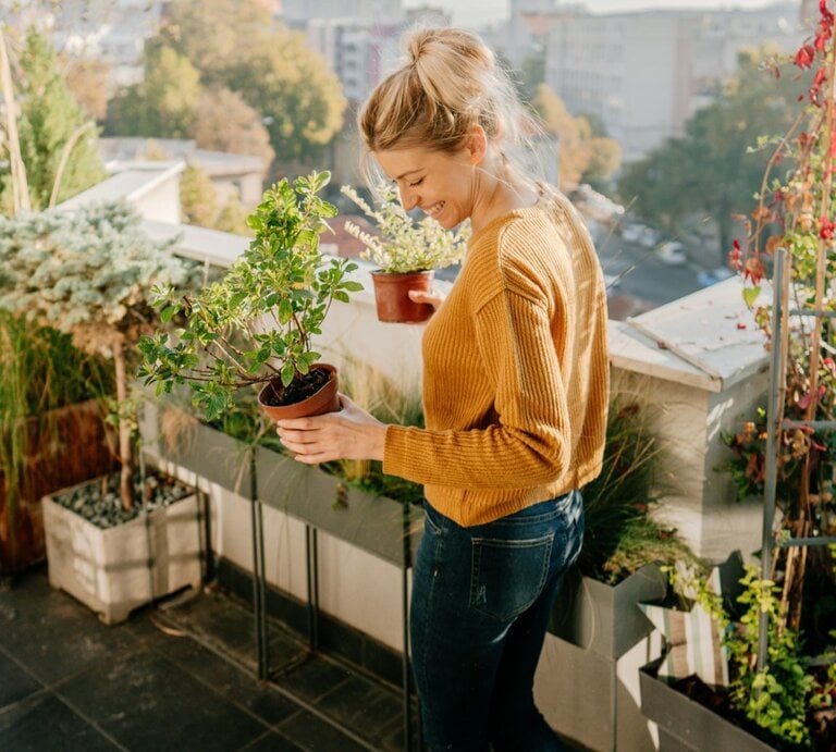 Eine Frau beim Urban Gardening auf ihrem Balkon mit Blumentöpfen und Gemüsepflanzen am Geländer und am Boden. | © gettyimages/ AleksandarNakic