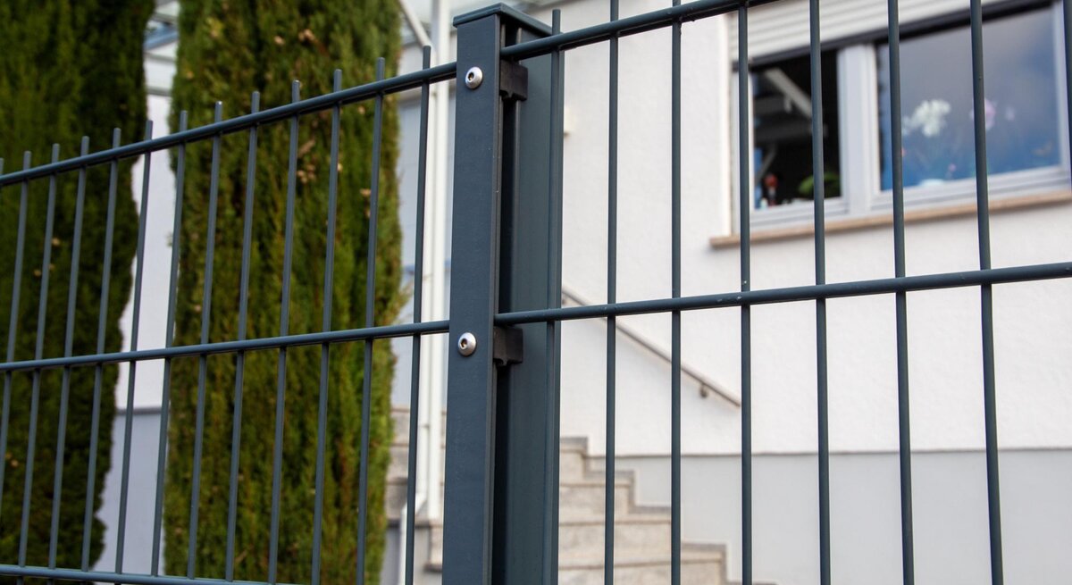 Stabmattenzaun in Nahaufnahme. Dahinter Bäume und ein Haus. 