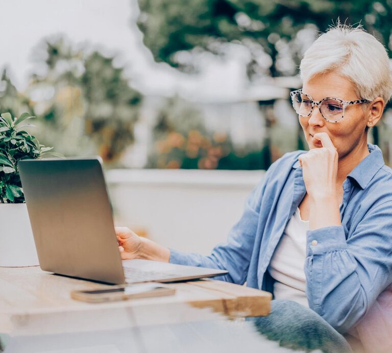 Frau mit markanter Brille zwischen 50 und 70 mit kurzen weiß-blonden Haaren sitzt draußen auf dem Balkon vor dem Laptop.  | © Getty Images