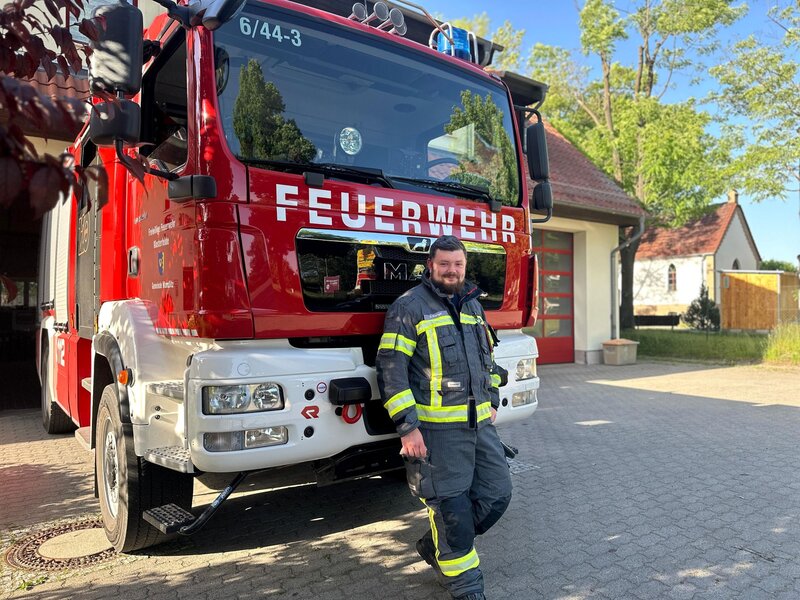 Ein junger Feuerwehrmann in Schutzkleidung lehnt lächelnd vor rotem Löschfahrzeug auf sonnigem Hof vor Wache. | © Adrian Prengel