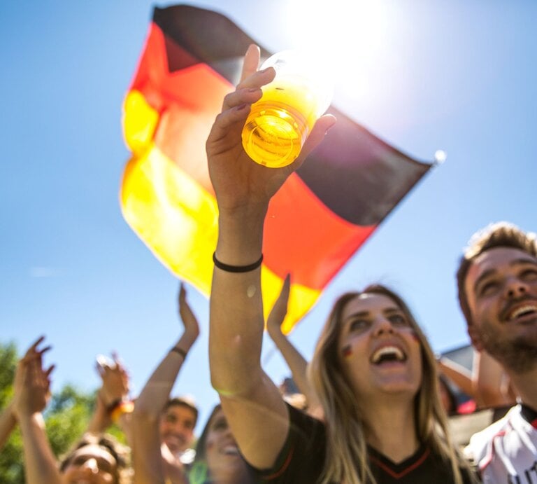 Jubelnde junge Fußballfans stoßen mit Bier an, schwenken eine Deutschlandflagge und feiern unter strahlend blauem Himmel. | © GettyImages/LeoPatrizi