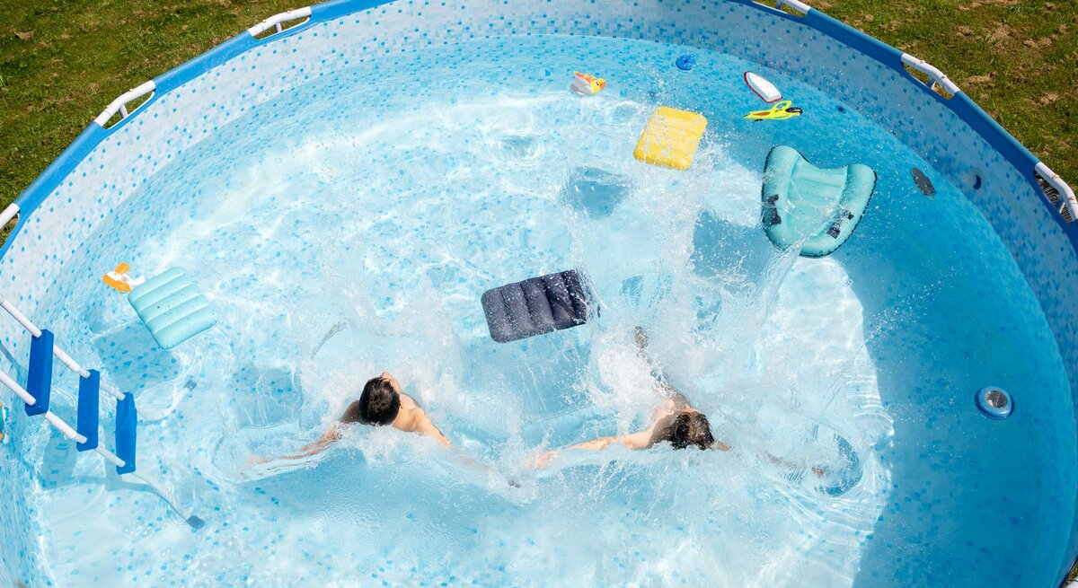 Zwei Kinder planschen ausgelassen in einem runden Gartenpool, während Wasserfontänen und bunte Schwimmhilfen die Szene beleben.