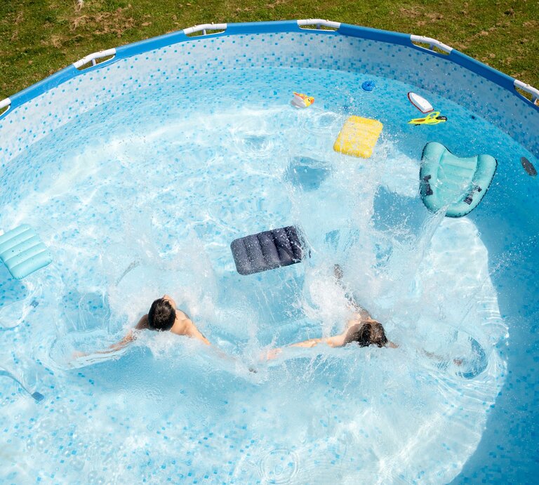 Zwei Kinder planschen ausgelassen in einem runden Gartenpool, während Wasserfontänen und bunte Schwimmhilfen die Szene beleben. | © GettyImages/Vladimir Vladimirov