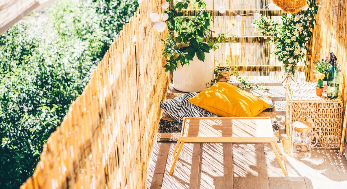 Sonniger Balkon mit Bambusverkleidung, gelbem Kissen, Pflanzen und Rattanmöbeln als gemütliche grüne Ruheoase.