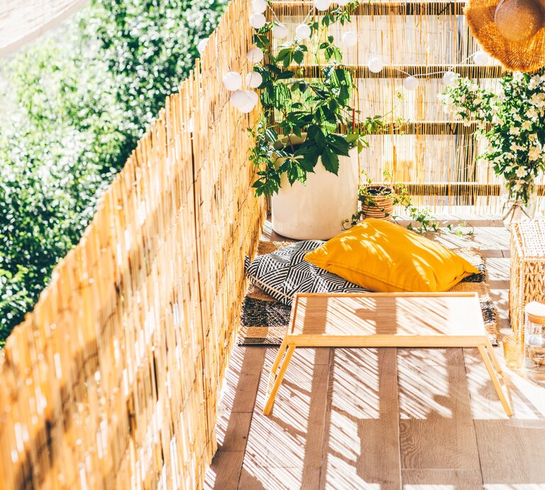 Sonniger Balkon mit Bambusverkleidung, gelbem Kissen, Pflanzen und Rattanmöbeln als gemütliche grüne Ruheoase. | © Lidl/Canva/Collage:bildderfrau.de