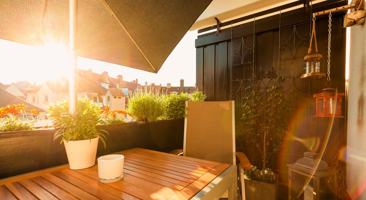 Sonniger Balkon mit Holztisch, beigem Stuhl und üppigen Topfpflanzen; warmes Abendlicht taucht die Szenerie in Gold.