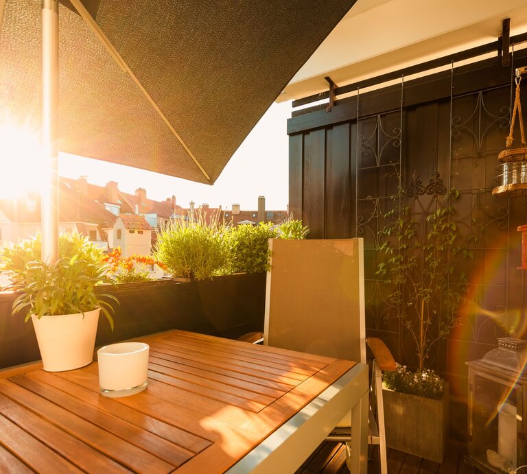 Sonniger Balkon mit Holztisch, beigem Stuhl und üppigen Topfpflanzen; warmes Abendlicht taucht die Szenerie in Gold. | © GettyImages/thethomsn