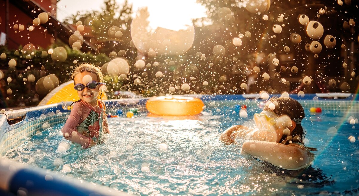 Zwei Kinder planschen und spielen mit Schwimmbrillen in einem Gartenpool an einem sonnigen Sommertag.
