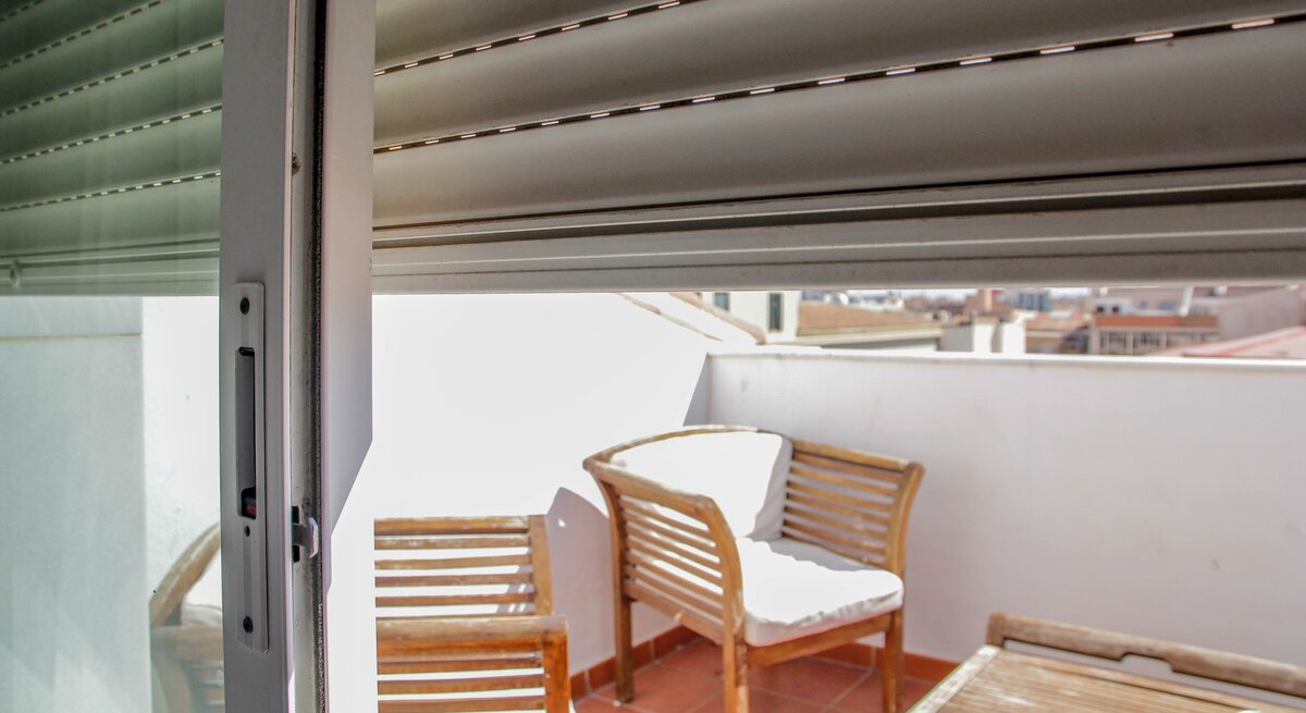 Ein Rollo ist halb heruntergelassen, eine Balkontür ist geöffnet und gibt den Blick auf eine Terrasse mit einem Tisch und einem Stuhl aus Holz mit weißem Bezug frei.