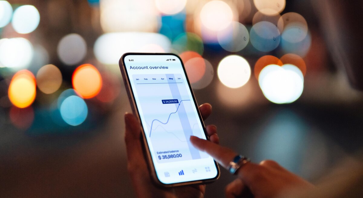 Frau guckt draußen auf ihr Smartphone. Darauf ist ein Aktienkurs zu sehen. 