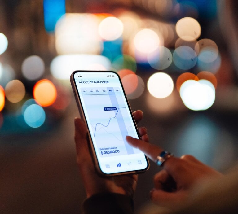 Frau guckt draußen auf ihr Smartphone. Darauf ist ein Aktienkurs zu sehen.  | © Getty Images